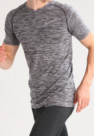 Mann in enganliegendem grauem, gemustertem kurzärmeligem Sportshirt und schwarzen, strukturierten Leggings läuft oder joggt vor schlichtem Hintergrund.