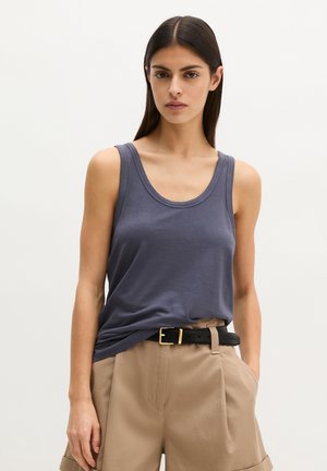 Vrouw met lang donker haar draagt een mouwloze donkergrijze tanktop, ingestopt in beige hoge taillebroek met zwarte riem, handen in de zakken.