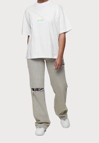 Osoba oblečená v bílém oversized tričku s malým zeleným a žlutým logem, světle šedých roztrhaných džínách a bílých teniskách, stojící na jednobarevném pozadí.