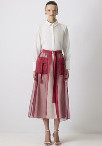 Femme portant une robe chemise blanche à manches longues avec une jupe superposée rouge transparente, dotée de grandes poches et d'une ceinture nouée à la taille.