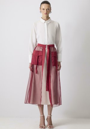 Donna che indossa un vestito camicia bianco a maniche lunghe con una gonna trasparente rossa sovrapposta, caratterizzata da grandi tasche e una cintura legata in vita.