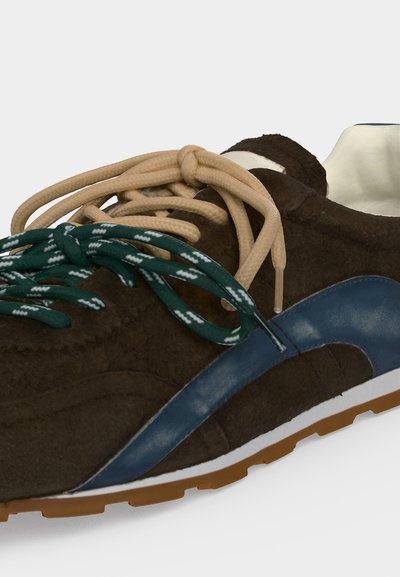 Baskets en daim marron avec un détail en cuir bleu et deux jeux de lacets, beige et vert avec des motifs blancs, sur fond blanc.