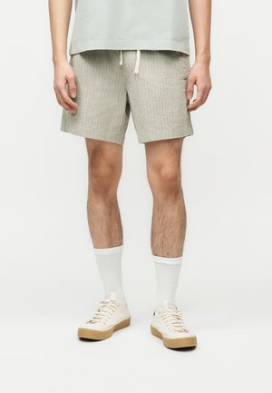 Personne portant un short en maille gris clair avec cordon de serrage, des chaussettes blanches montantes et des baskets écrues à semelles gomme, debout devant un fond pâle.