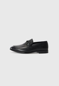 Mocassins en cuir noir avec une forme épurée, présentant un dessus texturé et une bride décorative avec une boucle en métal. Talon plat, design minimaliste.