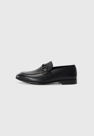 Mocasines de piel negra con una forma estilizada, que presentan una parte superior texturizada y una correa decorativa con una hebilla de metal. Tacón plano, diseño minimalista.