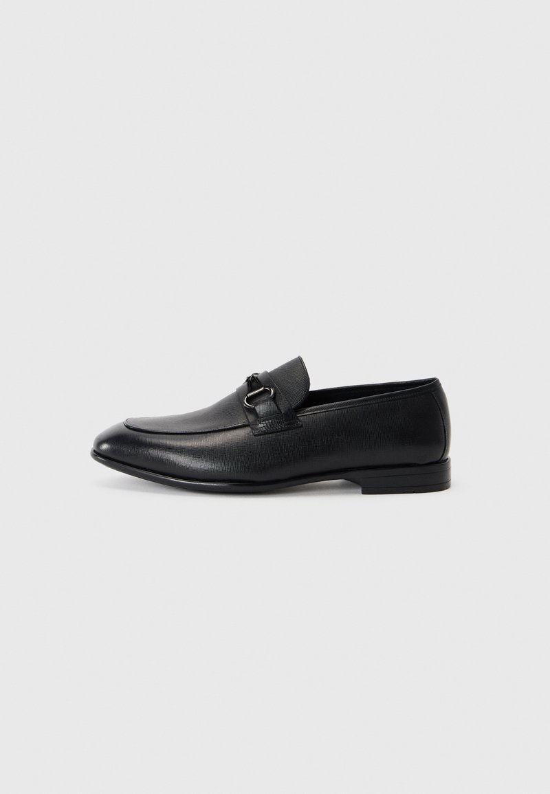 Mocassins en cuir noir avec une forme épurée, présentant un dessus texturé et une bride décorative avec une boucle en métal. Talon plat, design minimaliste.