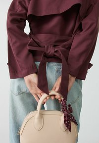 Bordeaux wikkeljack met strik in de taille, gecombineerd met lichtblauwe jeans en een beige handtas met een bordeaux gekleed sjaaltje als accent.
