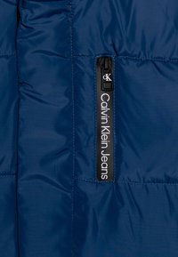 Granatowa kurtka puchowa z błyszczącego materiału, pionowa kieszeń na zamek oraz biała metka "Calvin Klein Jeans" po lewej stronie.