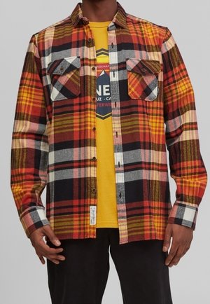 Mand iført en åben rød, gul og sort ternet skjorte over en gul grafisk t-shirt og sorte bukser, stående med armene afslappede.