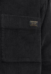 Camisa de pana negra con costillas verticales, un bolsillo rectangular en el pecho y una etiqueta de marca con letras doradas. Acabado texturizado y suave.