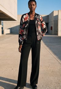 Frau steht im Freien und trägt eine schwarze Bomberjacke mit Blumenmuster, schwarzes Top, weit geschnittene schwarze Hose und schwarze Schuhe vor einem Betonhintergrund.