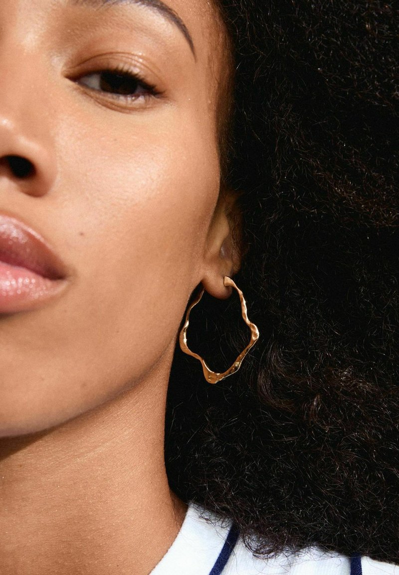 Pendiente de aro ondulado en oro con superficie texturizada. El cabello es rizado y oscuro. El tono de piel es liso y uniforme, con un maquillaje natural en los labios.