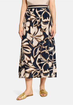 Femme portant une jupe mi-longue avec de grands motifs floraux beige et marron sur fond foncé et des sandales dorées à enfiler.