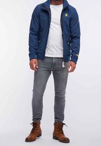 Giacca a zip blu navy con polsini a costine, con logo sul petto, indossata sopra una maglietta bianca e abbinata a jeans grigi e stivali marroni.