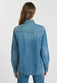 Vuxen som bär en medelblå denimskjorta och jeans, visad bakifrån mot en enkel ljus bakgrund.