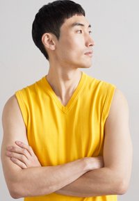 Yellow sleeveless V-neck shirt, smooth texture, fitted design. Model has arms crossed, showcasing the fabric's bright color against a neutral background.