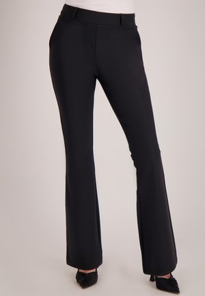 Vrouw die zwarte uitlopende broek en zwarte puntige hakken draagt, staand tegen een effen lichte achtergrond.