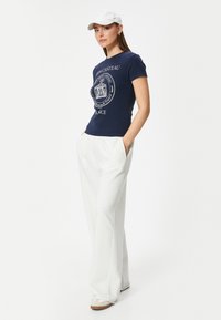 Marineblå kortærmet t-shirt med hvid grafisk tryk, parret med hvide brede bukser og beige sko, kompletteret med en hvid kasket.