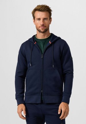 Man met kort haar en baard, die een donkerblauwe hoodie met ritssluiting en voorzakken draagt en daaronder een groen overhemd, tegen een effen achtergrond.