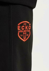 Svarte joggebukser med brodert oransje skjoldlogo som viser "FC ECKŌ UNLIMITED" og "1993." Myk tekstur med synlige sidesømmer.