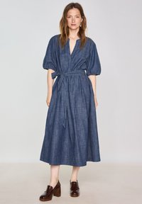 Abito midi in denim con maniche a sbuffo, scollatura a V profonda, fascia di tessuto in vita e vestibilità rilassata in blu scuro. Abbinato a scarpe con plateau marroni.