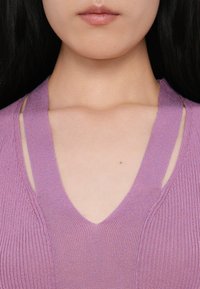Gros plan du bas du visage et du cou d'une femme portant un haut en maille violet avec un col en V et des découpes aux épaules.