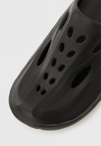 Fekete lábbeli texturált felülettel, több szellőzőnyílással, sima lekerekített élekkel és rugalmas kialakítással a kényelem érdekében.