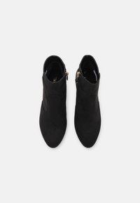 Un par de botines de ante negro con puntas redondeadas y cremalleras doradas en los laterales, vistos desde arriba sobre un fondo blanco.