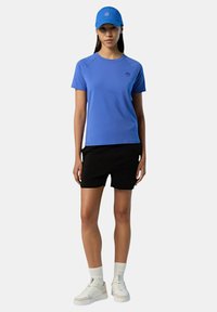 Camicia azzurra a maniche corte con logo, pantaloni neri, scarpe bianche e un cappellino blu. Design moderno con una vestibilità aderente, tessuto morbido e look casual.