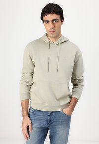 Hoodie cinza claro feito de tecido macio, com um bolso frontal, cordões no pescoço e um pequeno logotipo no peito. Usado com jeans azuis.