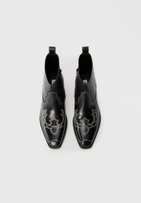 Botas de tobillo de cuero negro con punteras afiladas, que presentan intrincadas costuras plateadas y un diseño asimétrico. Cremalleras laterales para un fácil uso.