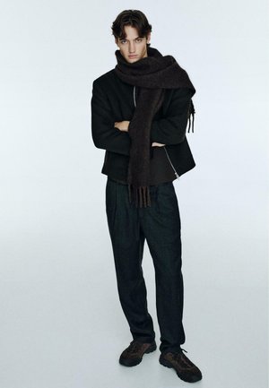 Brown wool scarf drapes over a black zip-up jacket, paired with dark trousers and brown sneakers. Model stands confidently with arms crossed.