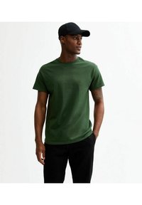 Grön bomullströja med rund hals, korta ärmar och avslappnad passform, ihop med svarta byxor och en svart keps.