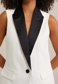 Kvinna klädd i en ärmlös vit blazer med svart satinrevers och en svart knapp, poserar med händerna bakom ryggen.