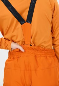 Pantalons de ski orange avec bretelles noires ajustables, présentant une taille froncée et une texture lisse. Une vue rapprochée met en avant les détails de couture.