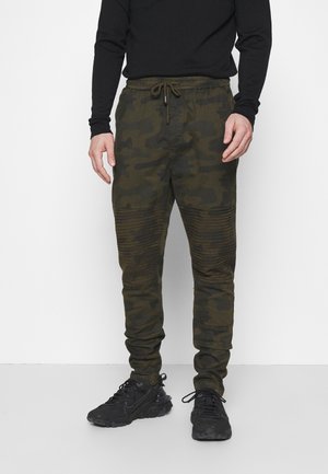 Man wearing dark green camouflage jogger pants with ribbed knee panels, black long-sleeve shirt, and black sneakers on a white background.
