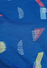 Tela azul con patrones geométricos en blanco, rojo y verde. Presenta círculos, triángulos y líneas de diferentes tamaños y orientaciones.