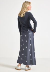 Femme blonde souriante, portant un haut à manches longues noir, une jupe maxi grise avec des étoiles blanches et des baskets blanches, se tenant devant un fond blanc.