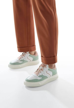 Sneakers in pelle bianca e suede verde presentano una robusta suola in gomma bianca, dettagli rosa e lacci in tessuto, abbinati a pantaloni marroni con risvolto.