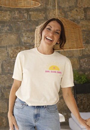 Lächelnde Frau mit braunen Haaren, die ein beiges T-Shirt mit der Aufschrift „Sun, Sand, Surf“ und hellblaue Jeans trägt, steht drinnen vor geflochtenen Lampen und einer Steinwand.