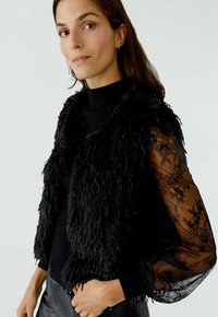 Schwarze Weste aus einem strukturierten, flauschigen Material, die über einem schwarzen Langarmoberteil mit durchsichtigen Spitzeärmeln und einer figurbetonten Silhouette getragen wird.