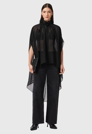 Kobieta ubrana w przezroczystą czarną bluzkę z wysokim kołnierzem i guzikami, szerokie czarne spodnie, czarne botki oraz trzymająca czarną kopertówkę.