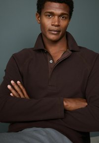 Polo de manga larga texturizada en marrón con cuello abotonado. Presenta costuras en contraste y una tapeta cosida. El modelo está posando con los brazos cruzados.