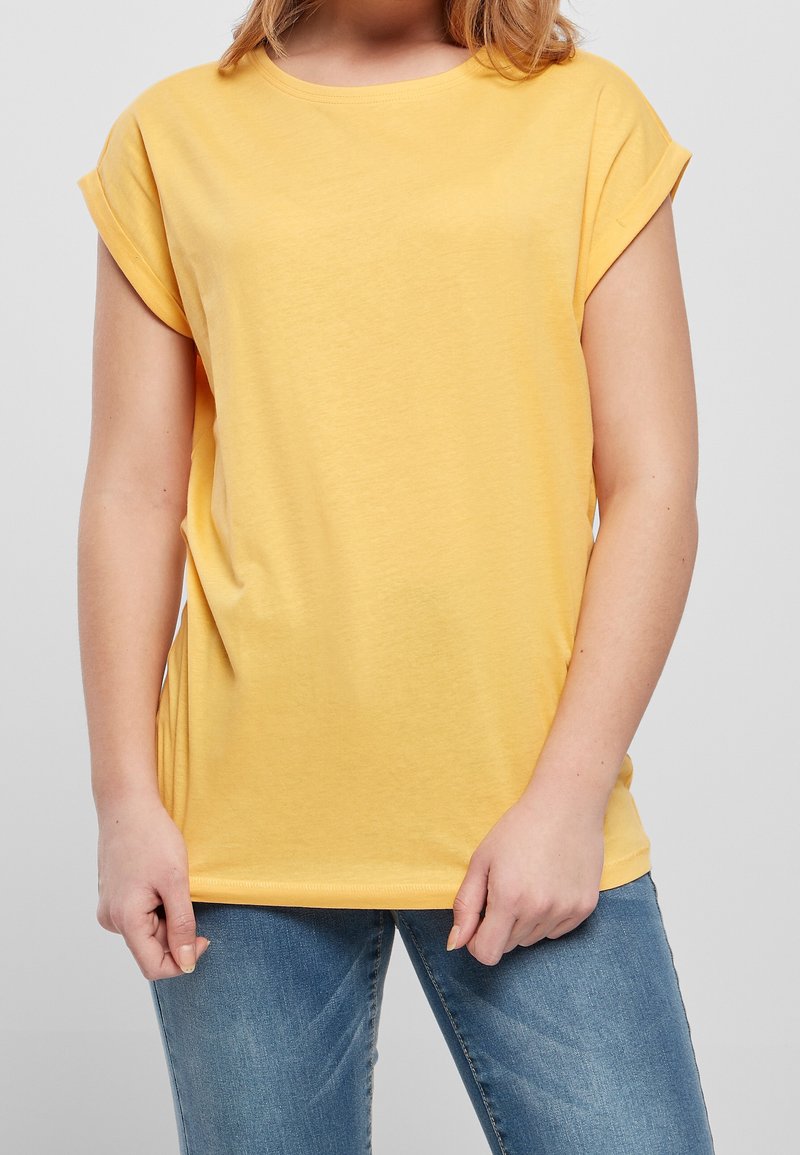 Personne portant un t-shirt jaune à manches courtes et un jean bleu, debout les mains près des hanches devant un fond uni.