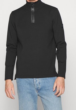 Mann trägt schwarzen Langarmpullover mit halbem Reißverschluss und hohem Kragen, kombiniert mit hellblauen Jeans, steht vor einem schlichten Hintergrund.