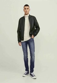 Mlad moški stoji, oblečen v črno bomber jakno, belo turtleneck pulover, modre kavbojke in črne visoke superge.