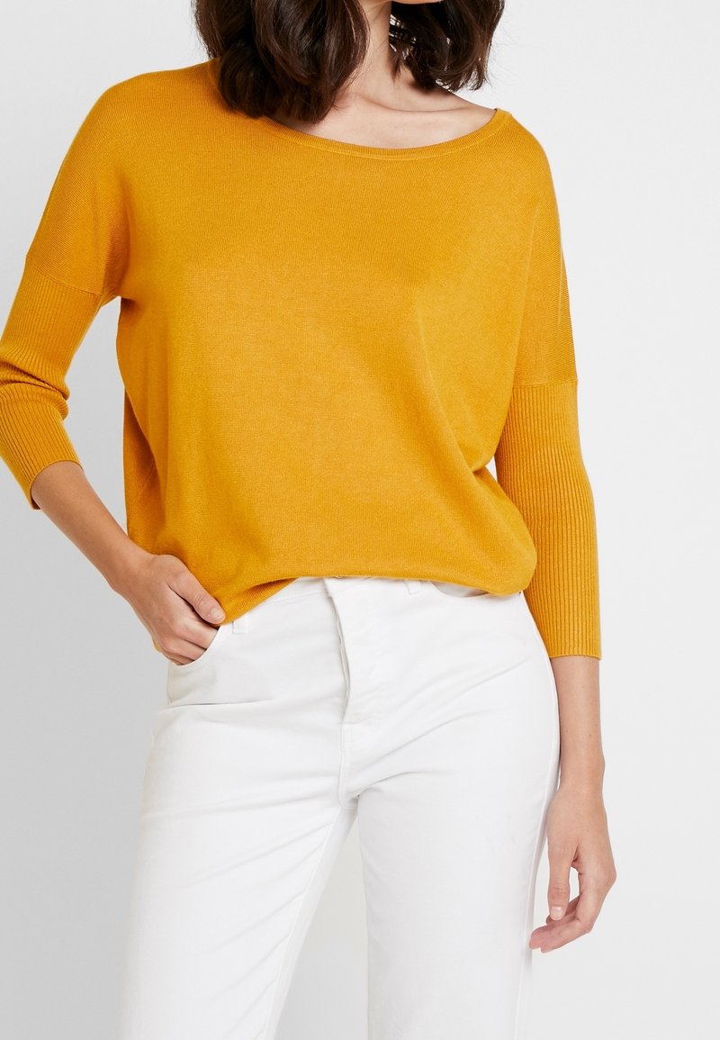 Femme portant un pull en maille jaune moutarde à manches longues avec des poignets côtelés et un pantalon blanc, main glissée dans la poche, sur un fond uni.