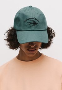 Zöld baseballsapka, amely anyagból készült, elöl hímzett "Hazafelé" felirattal és madármintával. Görbült perem, hatpaneles szerkezet.