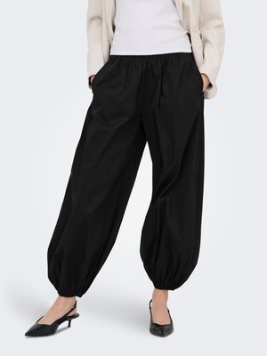 Rennot mustat housut, joissa joustavat lahkeet, mustien slingback-korkojen, valkoisen topin ja beigen bleiserin kanssa, malli seisoo kädet taskuissa.