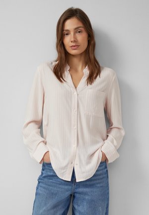 Junge Frau mit braunen Haaren, die ein hellrosa Hemd mit Knöpfen und blaue Jeans trägt, steht mit den Händen in den Taschen vor einer grauen Wand.